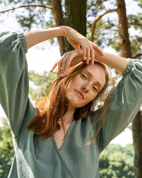 A fair-skinned person with reddish-brown hair and freckles on their face looks towards the camera, with their arms raised above their head. They are wearing a mint green v-neck garment with puffed sleeves. The background is blurred but appears to be trees and greenery.