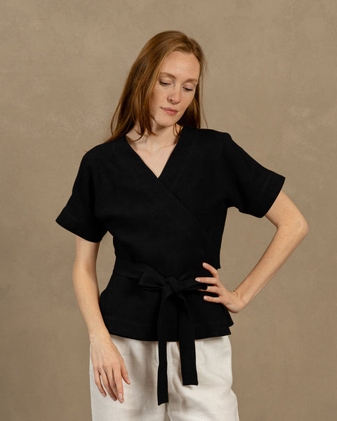 A fair-skinned person with long, auburn hair, wearing black V-neck linen wrap top with short, wide sleeves and light-colored pants. They are holding the ends of the top's tie in front of them, forming a bow.