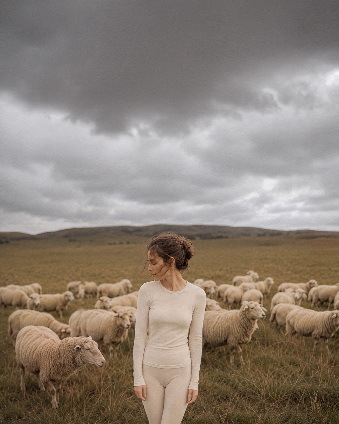 A woman in a cream-colored long-sleeve bodysuit stands in a grassy field surrounded by sheep under a dramatic, cloudy sky.