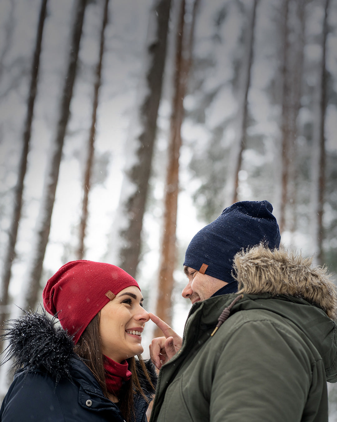 Smiling couple wearing winter hats and jackets, standing close together in a snowy forest