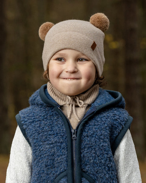 A young child with light brown curly hair and a subtle smile, is wearing a beige knit hat with brown pom-pom ears, a beige turtleneck, a navy blue fuzzy zippered vest, and a light-colored long-sleeved shirt underneath. The background is a blurred forest.