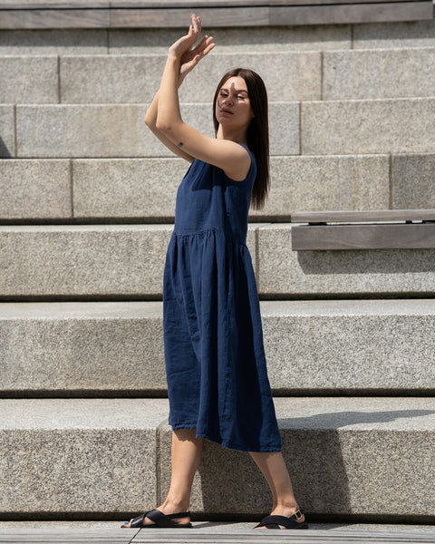 Young woman posing in a city, wearing linen smock dress Maya in a Storm blue cover. Woman posing with her hands up in the air, to cover sunshine.