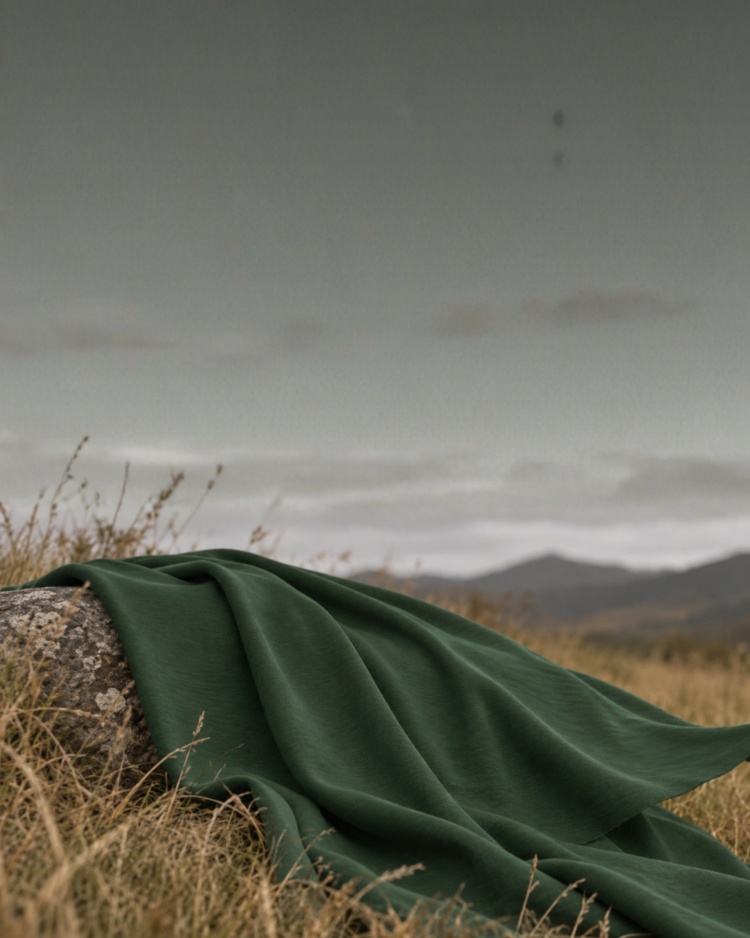 A piece of textured dark green fabric is draped over a weathered rock in a field of dry, wild grass. The fabric's edge catches a gentle breeze against a softly blurred backdrop of rolling mountains and a hazy, overcast sky.