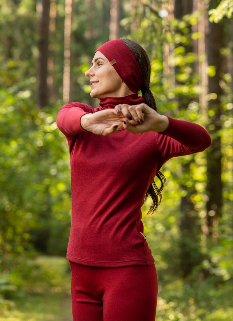 Woman stretching outdoors wearing red merino wool base layer and headband, breathable thermal activewear for hiking, running, and outdoor fitness in changing weather conditions.