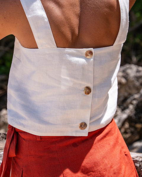 A close-up shot from behind shows a woman wearing a pure white sleeveless crop top with three round, light brown button closures down the center back. The linen top has wide straps and is paired with a cinnamon red linen wrap skirt tied at the side. The background is slightly blurred, suggesting an outdoor setting with natural elements like wood and foliage.