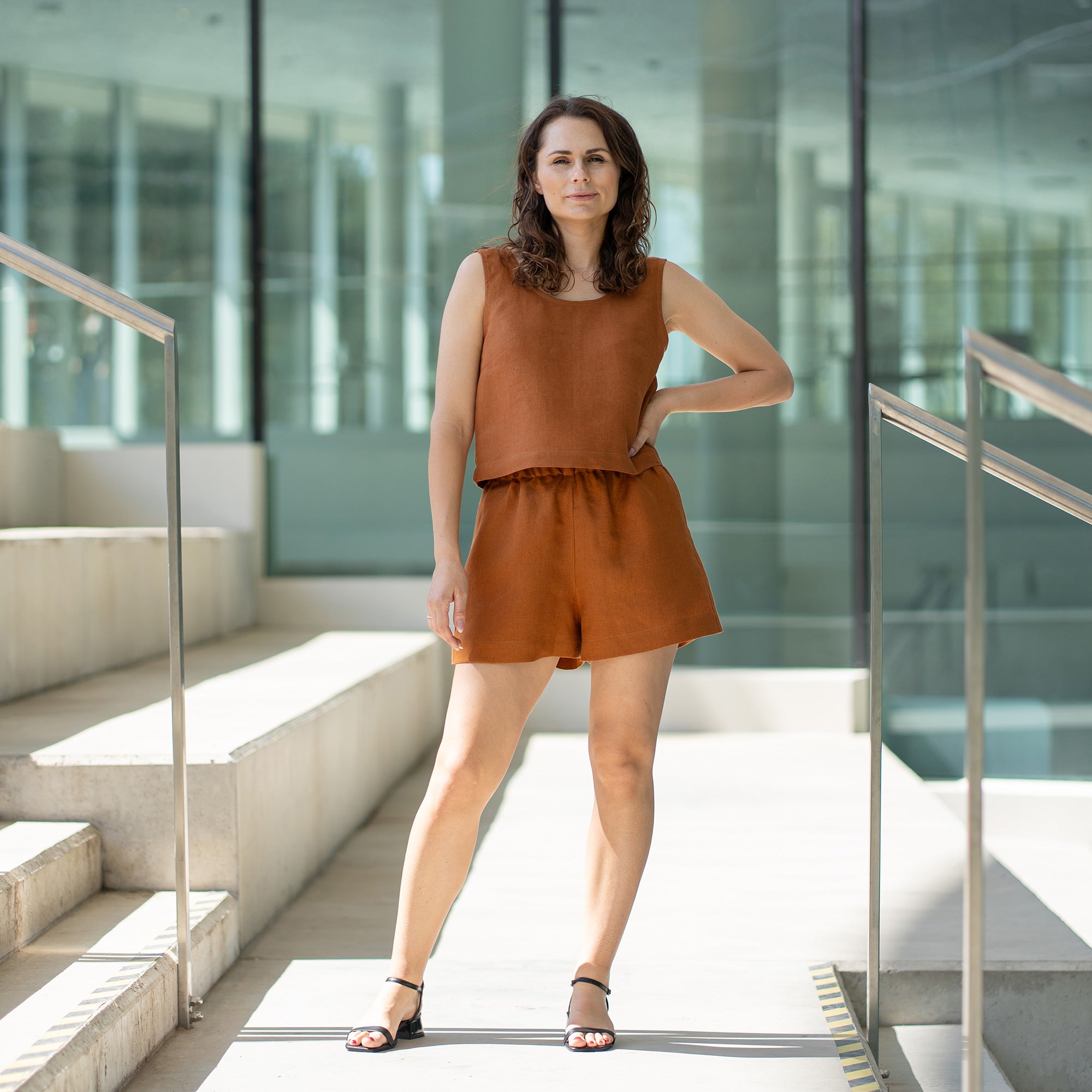 A woman with brown hair, wearing a matching almond brown sleeveless top and shorts, looks over her shoulder while standing on outdoor concrete stairs with metal railings.