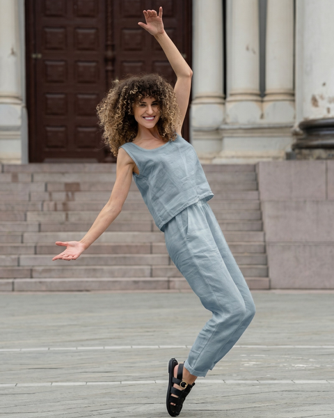 A woman with curly hair smiling brightly while posing playfully in a stone plaza, leaning back slightly with her arms gracefully outstretched and one leg lifted. She is wearing a light dusty blue linen set consisting of a sleeveless top and cropped pants.