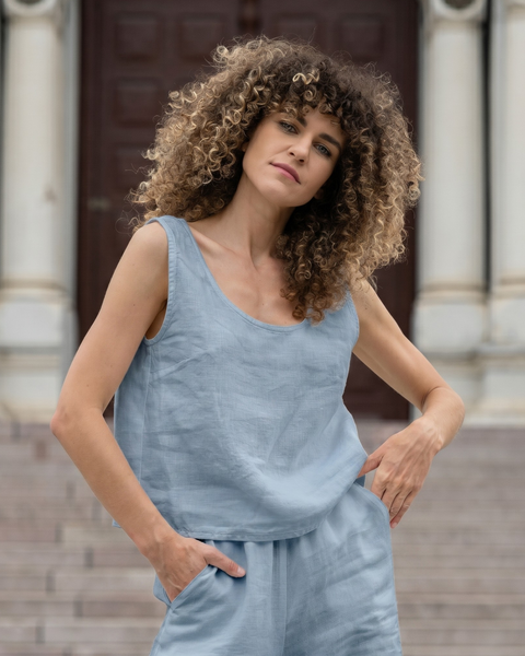 A woman with curly hair standing confidently in front of grand stone steps, with her hands in the pockets of her light dusty blue linen pants. She is wearing a matching sleeveless linen top and looking directly at the camera with a calm expression.