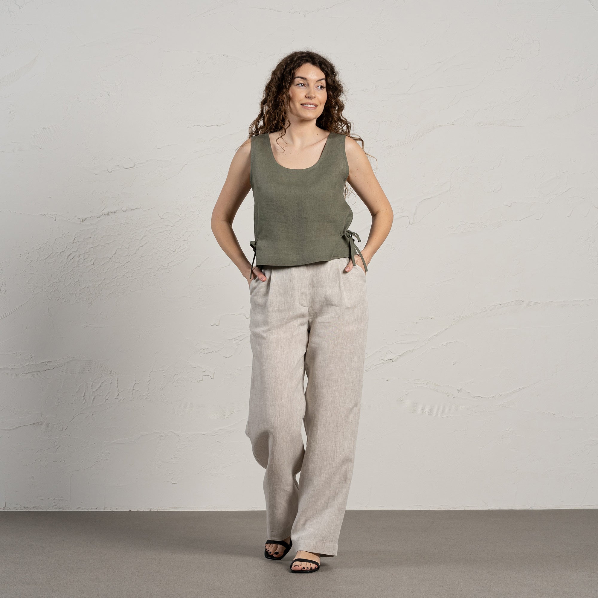 A smiling woman with long, curly brown hair wears a sleeveless linen crop top with side ties and light-colored wide-leg linen pants. She stands in front of a textured light gray wall, with one hand in her pocket.