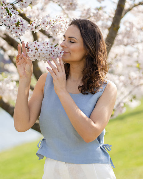 A woman with brown, wavy hair, wearing a cloudy blue sleeveless top, stands with her eyes closed, gently bringing a branch of white cherry blossoms to her nose as if to smell them. A light blue body of water and green grass are visible in the soft background.