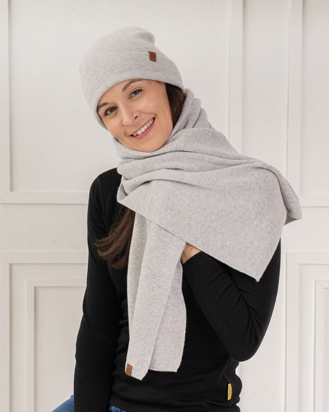 A smiling woman looking towards the camera, wearing a matching set of a light grey knit beanie and a light grey knit scarf wrapped generously around her neck. She wears a black long-sleeved top against a background of a bright white wall with classic paneling.