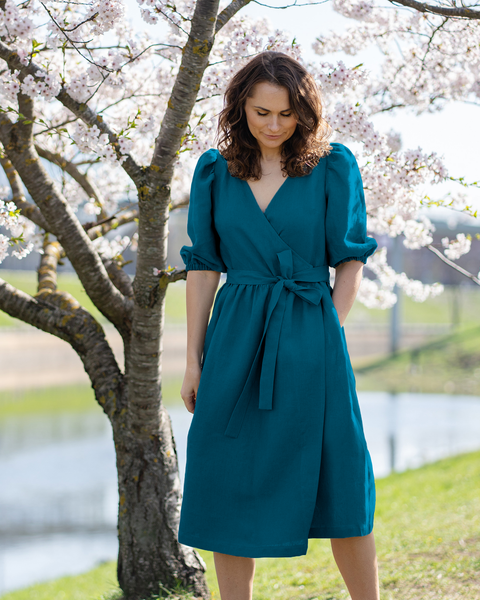 A light-skinned person with shoulder-length wavy brown hair is looking down with a gentle expression. They are wearing a menique linen wrap dress with short puff sleeves and a tied waist, with their hands in the side pockets. They are standing near a blossoming tree with white flowers, and a blurred background suggests a park-like setting with water and greenery.
