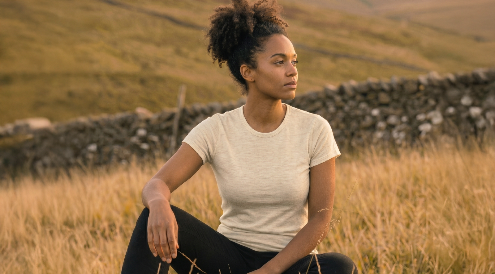 A woman with dark curly hair sitting in tall, dry grass during golden hour. She is wearing a cream-colored short-sleeved t-shirt and black pants, resting her arm on her knee while looking off to the side.