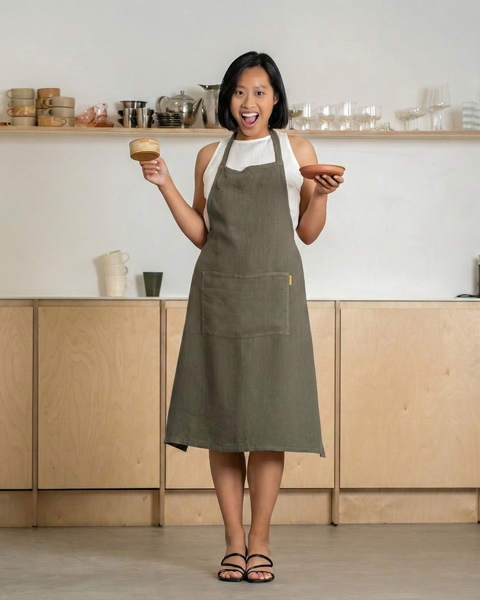Woman in a stone green linen apron holding a cup and plate, smiling in a minimalist kitchen interior.