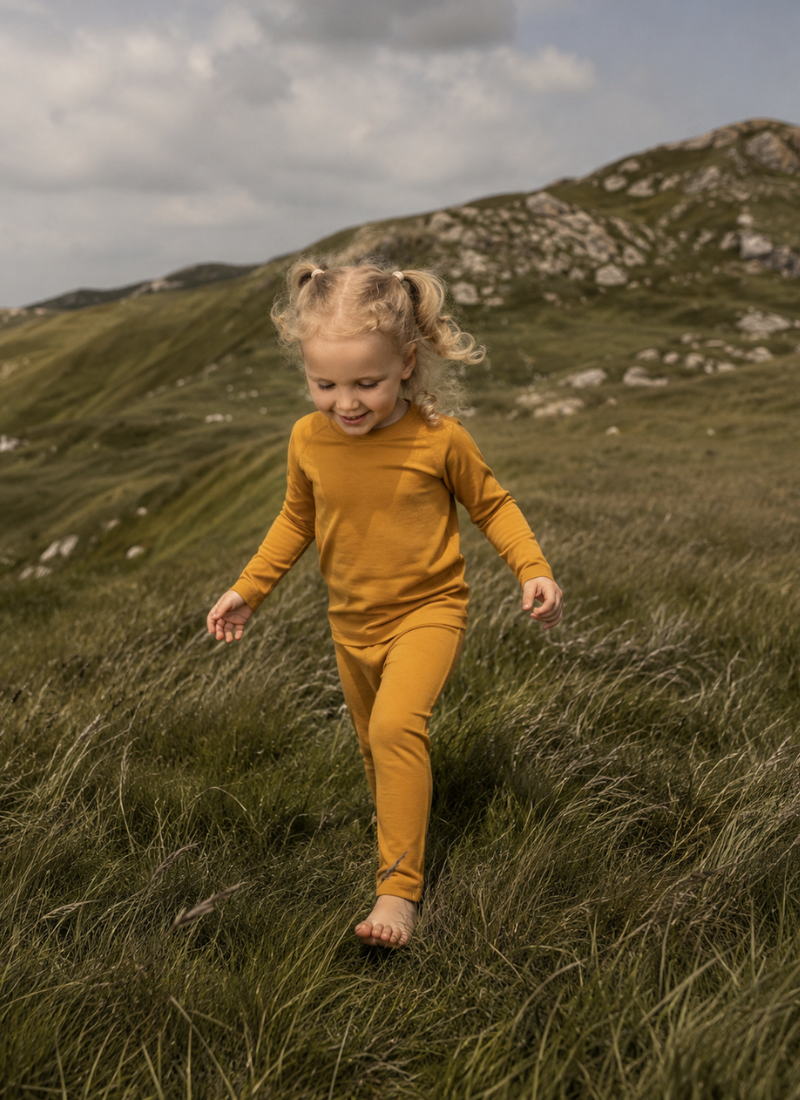 A young child with blonde pigtails runs barefoot through tall grass on a grassy hillside, wearing a matching mustard yellow long-sleeve top and pants set.