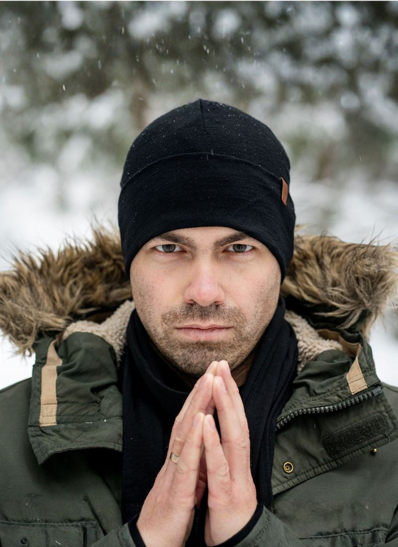 Man wearing a black beanie and green winter coat with a fur-lined hood, standing in a snowy forest.