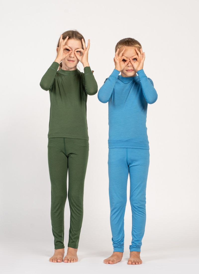 Two kids, a boy and a girl, wearing merino wool base layers, one in light blue and the other in dark green color. They are standing, making playful hand gestures on their face.