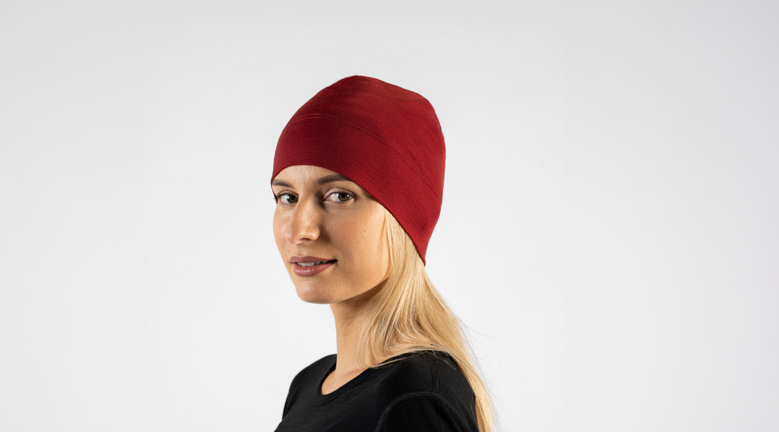 A blonde woman with a gentle smile looks towards the camera against a plain light grey studio background. She is wearing a snug, deep red knit beanie and a simple black crew-neck top.