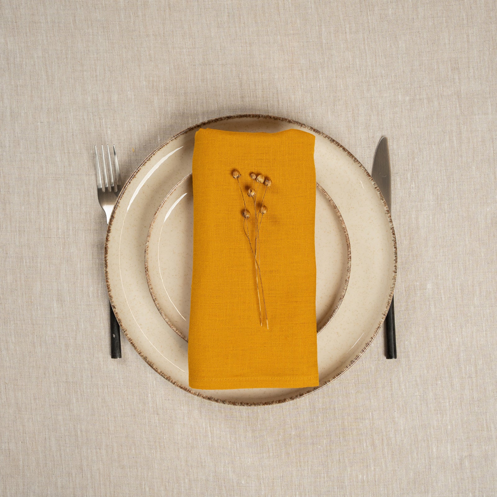 A table covered with a linen tablecloth, with a vase and plates on top of it. On top of the stacked plates is a Linen Napkin in Spicy Yellow color.