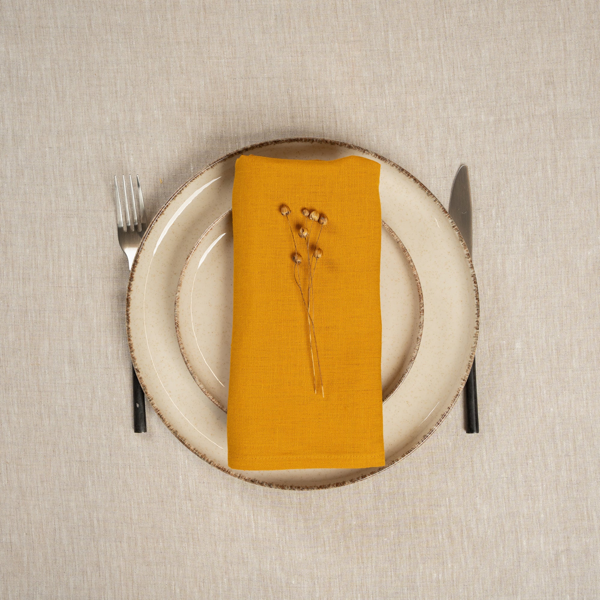 A table covered with a linen tablecloth, with a vase and plates on top of it. On top of the stacked plates is a Linen Napkin in Spicy Yellow color.