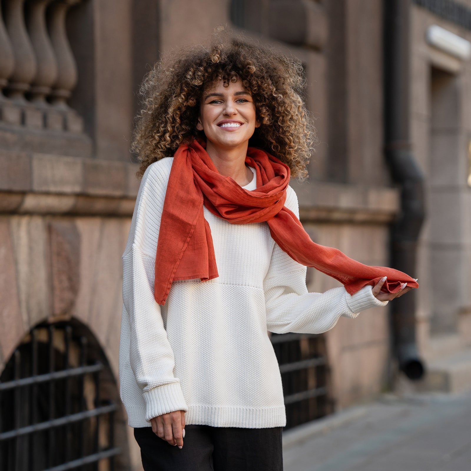 A woman wearing a checkered outfit and an orange scarf poses with their hands on the scarf.


