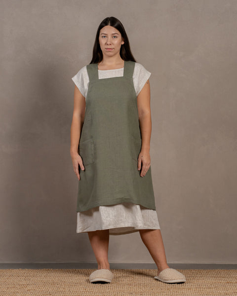A young woman with dark hair standing in a studio background, with her hands by her sides. She is wearing a linen dress with a linen cross-back japanese apron in stone green color in size L/XL on top.