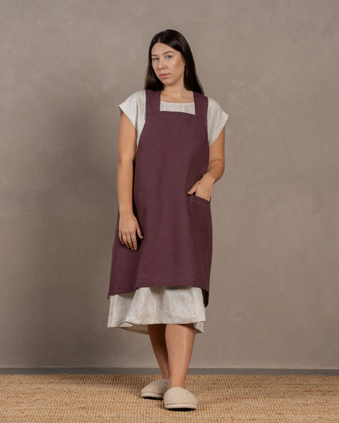 A young woman with dark hair standing in a studio background, with one hand inside the front pocket. She is wearing a linen dress with a linen cross-back japanese apron in shadow purple color in size L/XL on top.
