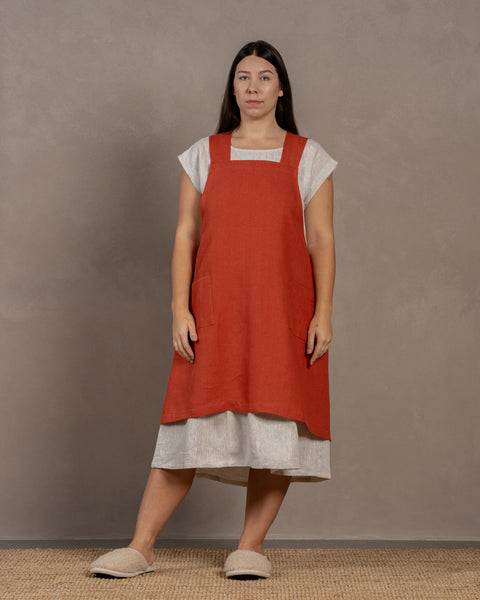 A young woman with dark hair standing in a studio background, with her hands by her sides. She is wearing a linen dress with a linen cross-back japanese apron in cinnamon red color in size L/XL on top.
