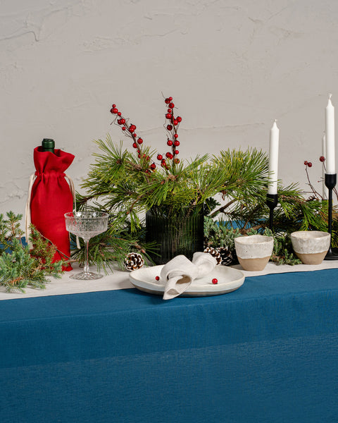 Festive holiday table setting with a deep blue linen tablecloth and a white linen table runner decorated with pine branches, red berries, pinecones, candles, and a red linen wine bag.