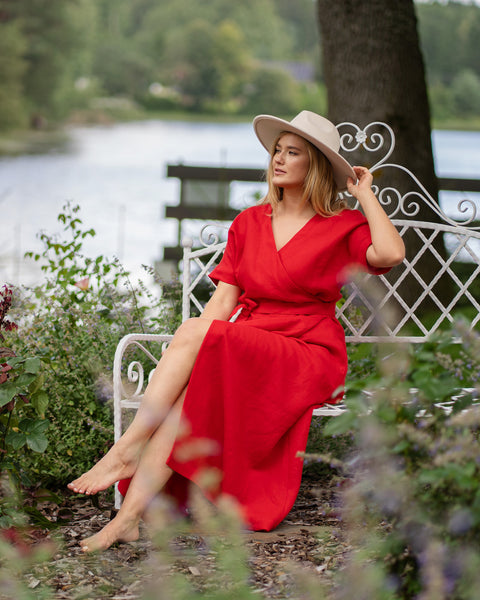 Woman wearing red wrap maxi dress and beige hat sitting on white garden bench near lake, flowing feminine summer dress