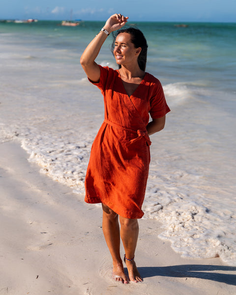 A woman with dark hair smiles and shields her eyes with one hand as she stands barefoot on a sandy beach. She is wearing a cinnamon red linen wrap dress with short sleeves. The ocean is in the background with small waves and a few boats visible in the distance under a bright sky.