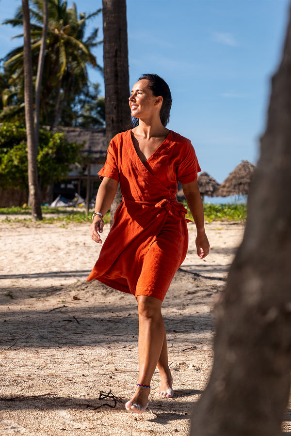 A smiling woman with long dark hair stands barefoot on a sandy beach, wearing a cinnamon nred linen sundress with thin straps. Her arms are outstretched to the sides. The ocean is in the background with gentle waves, and several boats are visible in the distance under a bright sky.

