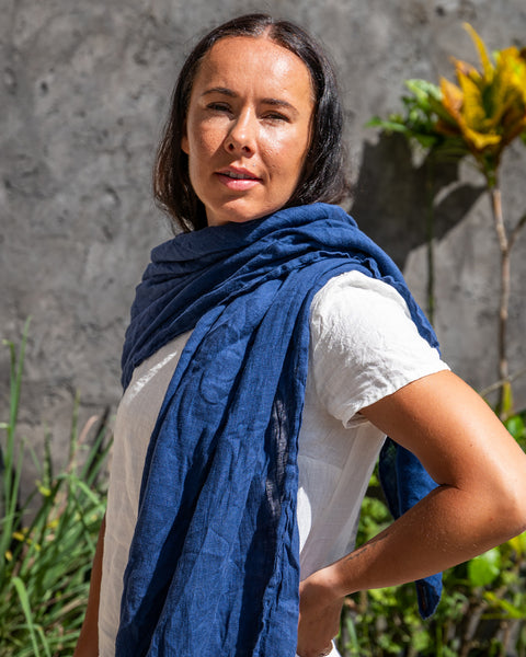 A person standing outdoors wearing a storm blue menique linen scarf draped over the shoulders, with a beige linen dress. The image captures a side profile of the person against a backdrop featuring a gray wall and greenery.