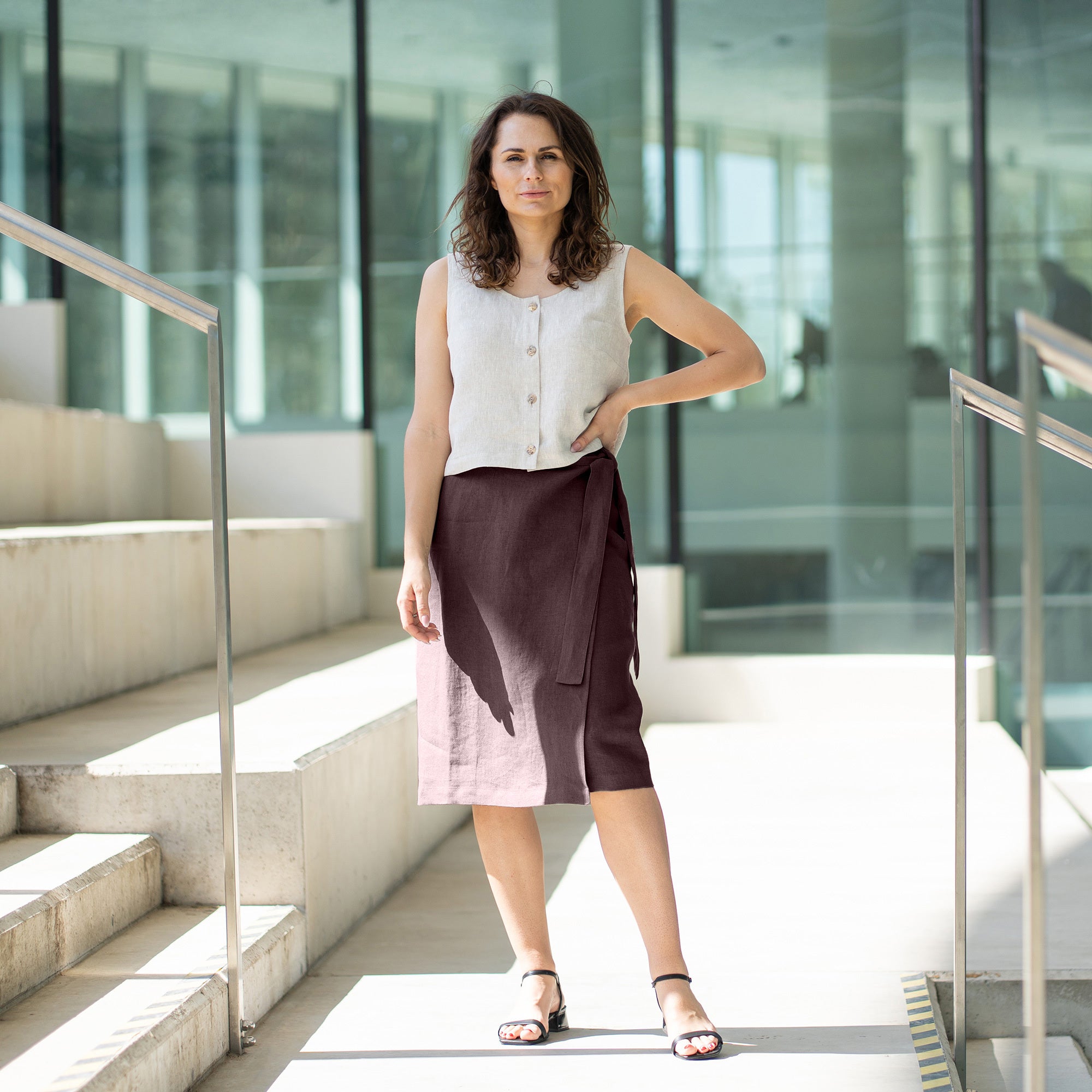 A woman with brown hair, wearing a natural linen button-front sleeveless top and a stone green wrap skirt, poses on outdoor concrete stairs.
