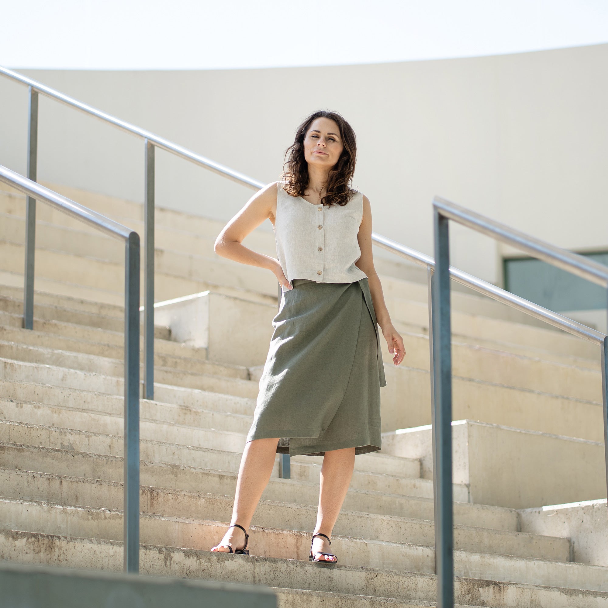 A person with short, curly dark hair stands in profile, looking down at their waist. They are wearing a white sleeveless top and a knee-length black wrap skirt with a long tie hanging down the front. On their feet are black sandals with multiple straps and a low block heel. To their left are two large potted plants against a light-colored wall.