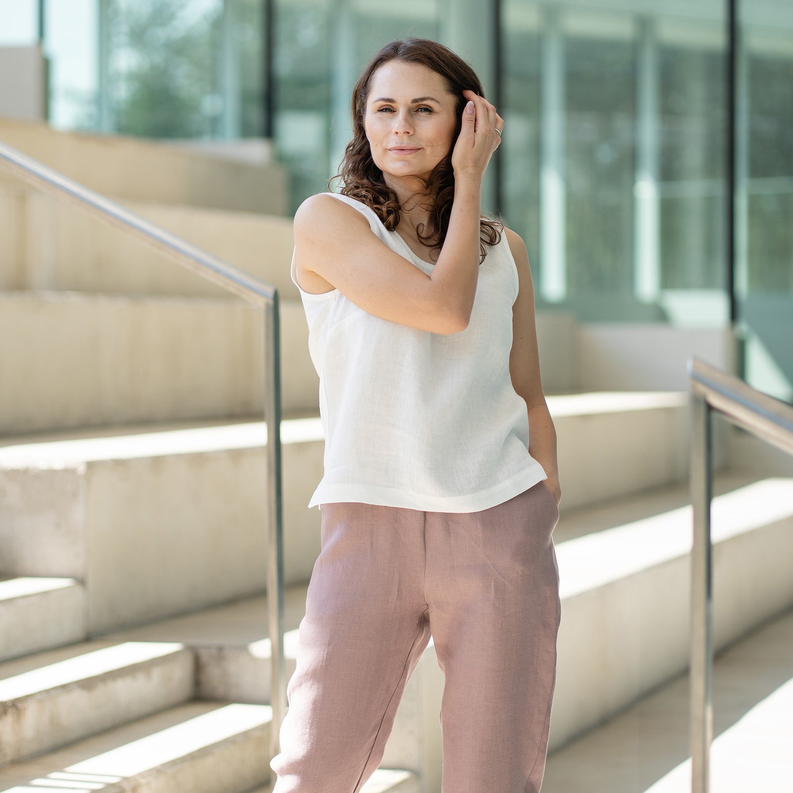 A woman with brown, shoulder-length hair, wearing a white sleeveless top and faded rose trousers, stands on concrete stairs inside a building with glass walls.