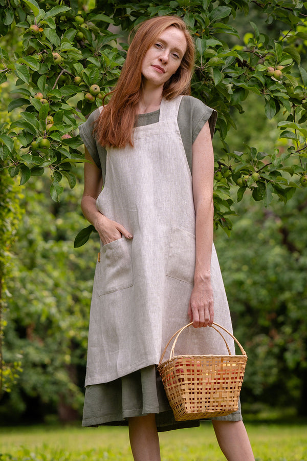 A woman with long red hair wearing a natural color linen apron, standing in a lush green garden and holding a woven basket.