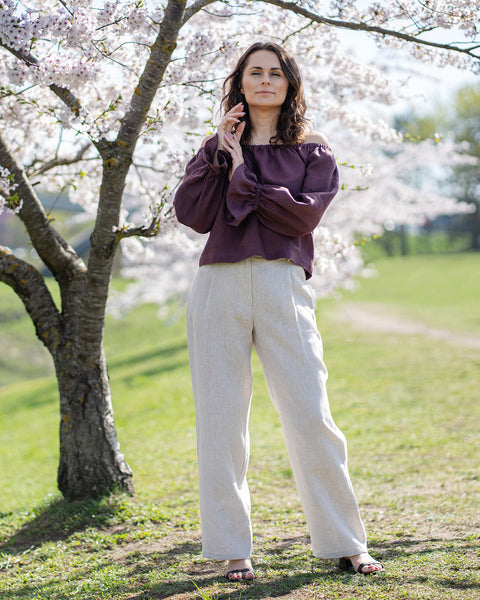 A woman with fair skin and dark, wavy hair, wearing a shadow puprle, off-the-shoulder top, is shown from the waist up.