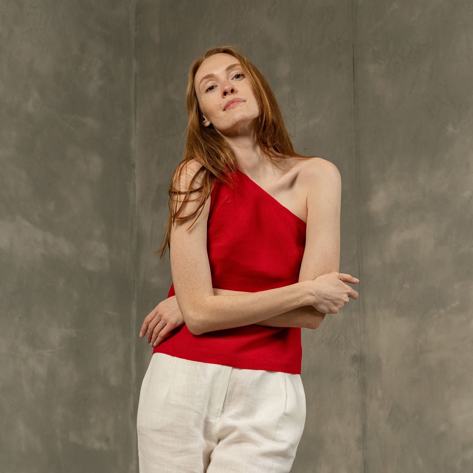 A woman with brown hair, wearing a teal blue one-shoulder top and wide-leg white trousers, walks down outdoor concrete stairs under a bright sky.
