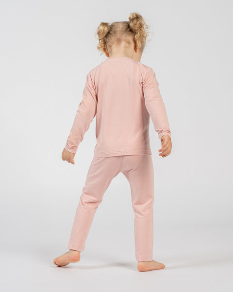 Child in pink long-sleeve top and matching pants, standing barefoot with back facing the camera and arms relaxed by the sides, showing the outfit from behind against a plain background.