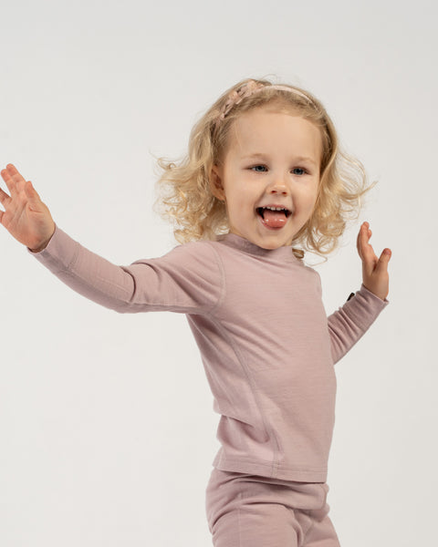 A playful child with curly blonde hair and a light pink floral headband stands against a white background, wearing a dusty pink long-sleeved top and matching pants. The child has their arms spread wide and their tongue slightly out, looking enthusiastically at the camera.