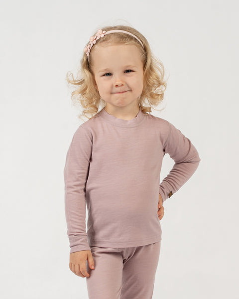 A child with curly blonde hair and a light pink floral headband stands against a white background, wearing a dusty pink long-sleeved top and matching pants. The child has one hand on their hip and is looking at the camera with a slight smile.