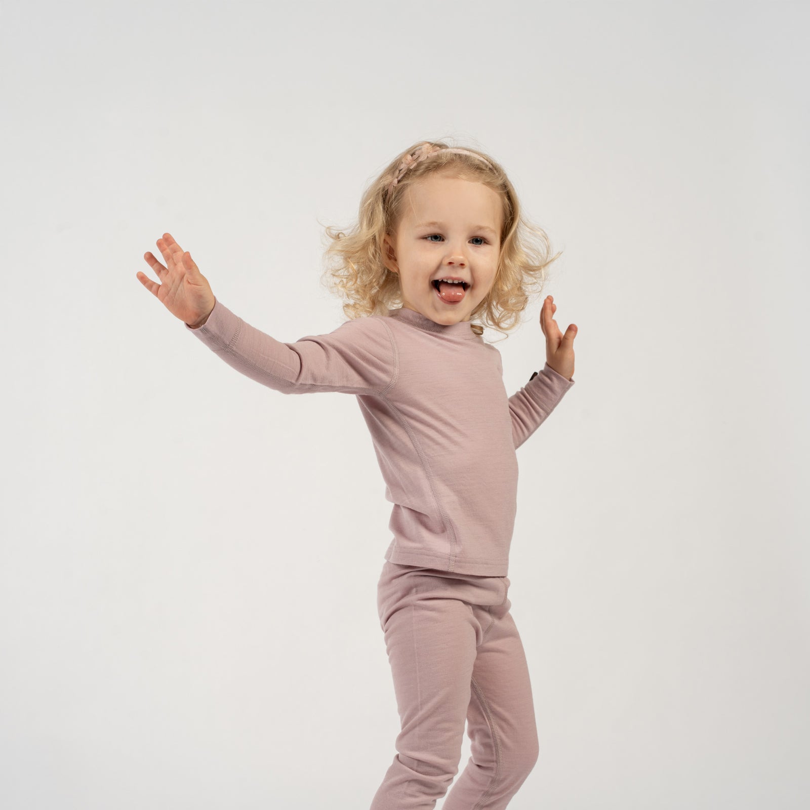 A child with blonde curly hair and a light pink floral headband stands against a white background, wearing a dusty pink long-sleeved top and matching pants. The child has one hand on their hip and is looking at the camera with a slight smile.
