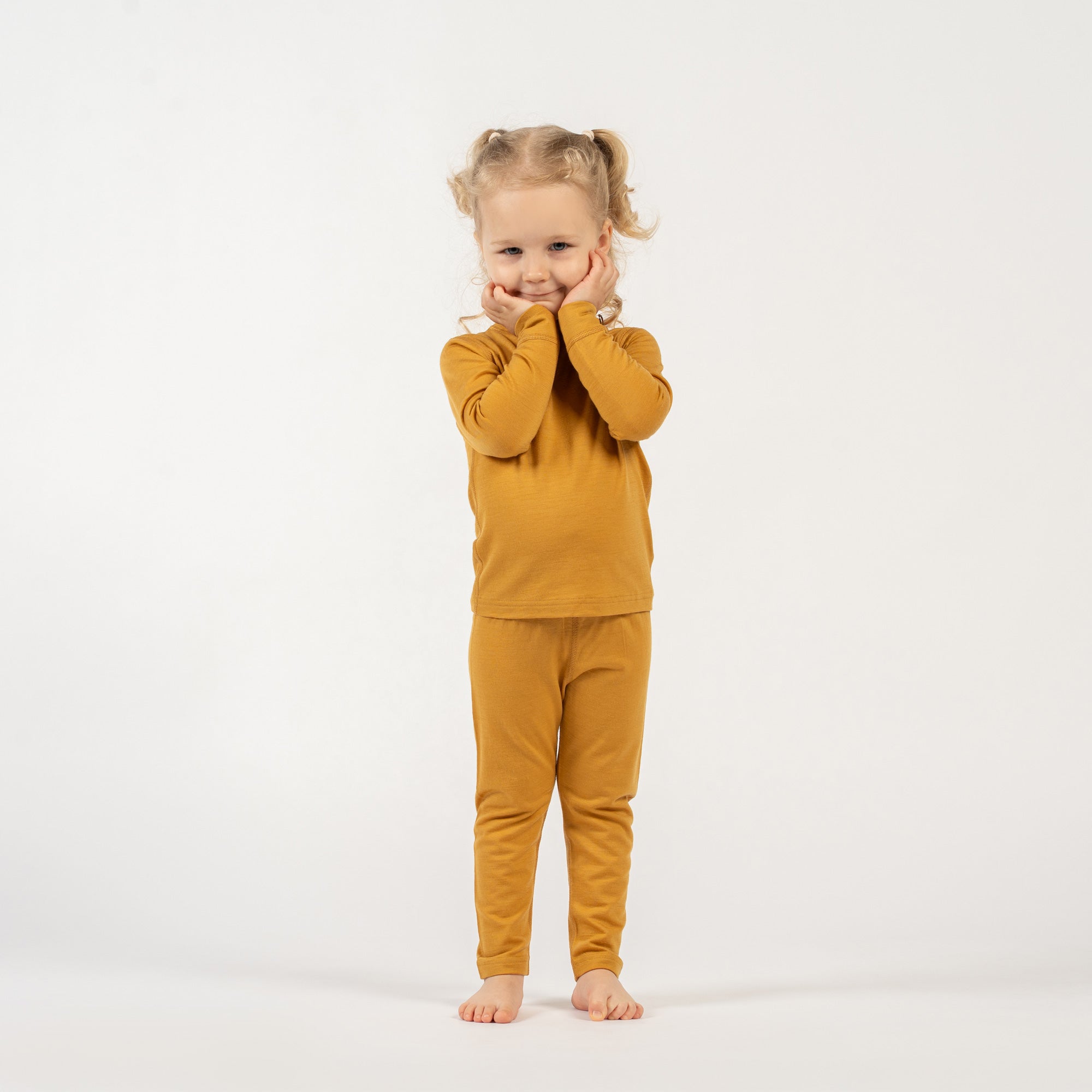 A young child with curly blonde pigtails, wearing a yellow long-sleeved top and matching pants, stands barefoot on a white background, looking directly at the camera with a gentle smile.