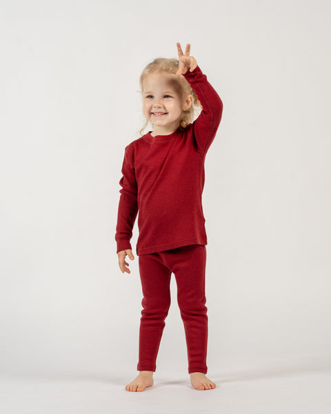 A young child with light curly hair stands facing forward, smiling broadly, and holding up both hands with two fingers extended on each, forming &quot;peace&quot; signs. The child is wearing a long-sleeved red top and matching red pants. The background is white.