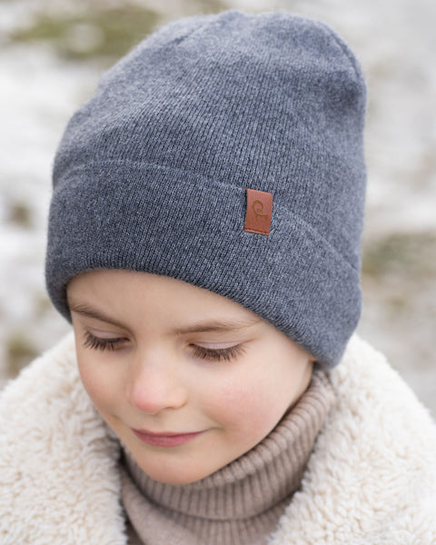 A close-up shot of a young child wearing a dark grey knit beanie with a brown label on the front, a beige turtleneck, and a cream-colored sherpa jacket. The child&#39;s eyes are cast downward, and the background is a blurred, lightly snowy outdoor scene.