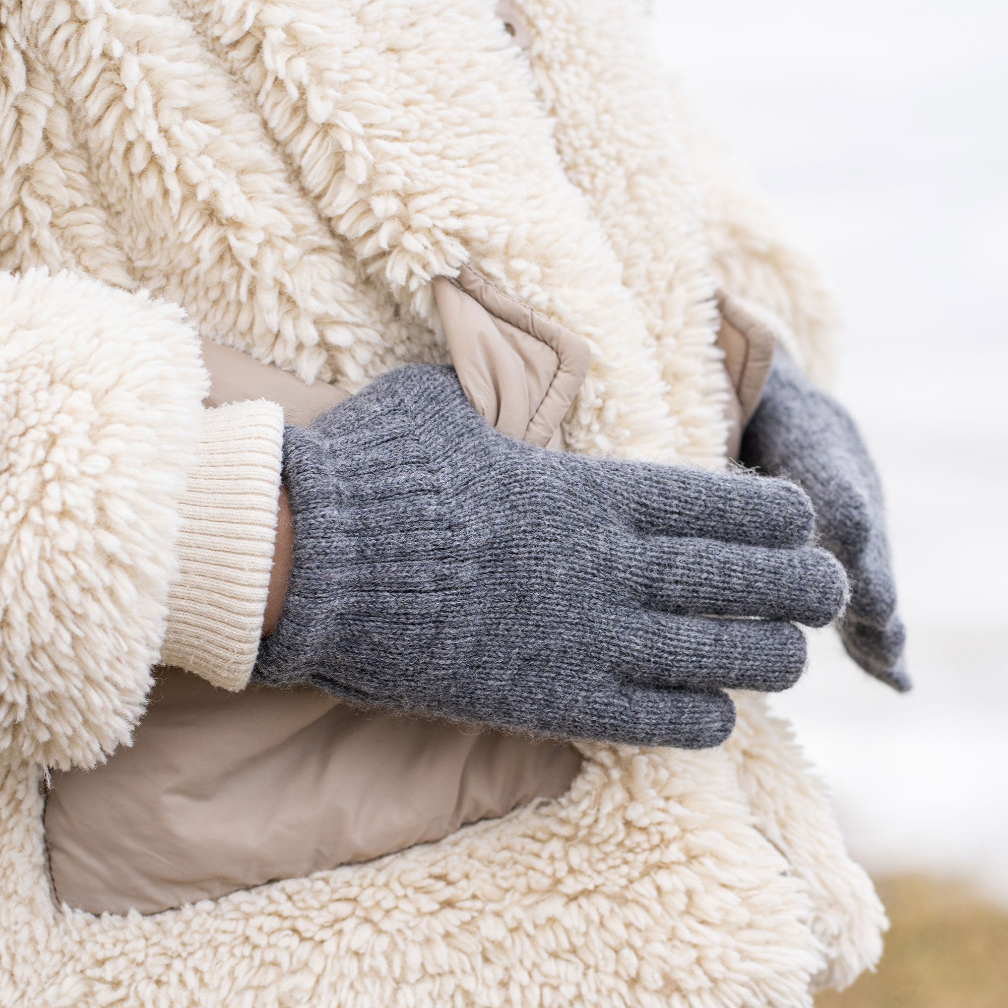 A hand wearing a light pink knitted glove with a ribbed wrist extends upwards and slightly to the left, revealing a plush, cream-colored sleeve on the arm, against a blurred light background.