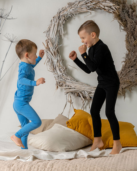 Two boys jumping on a bed, one of them is wearing the menique 160 kids&#39; set of long sleeve  crew and pants in light blue and the other in black color. They are play-fighting.