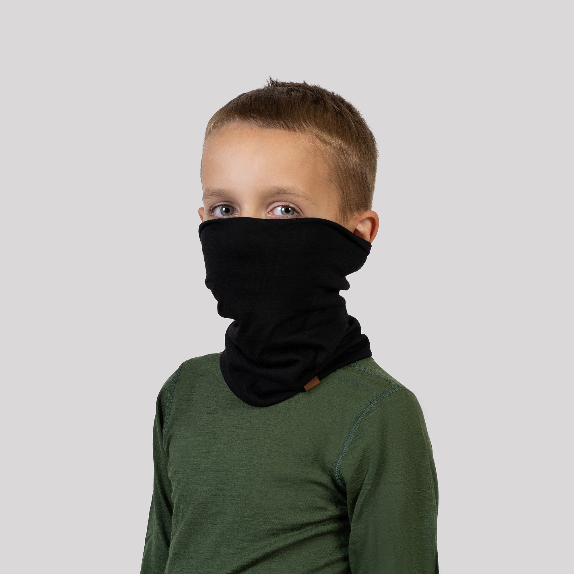 A cheerful child with blonde pigtails looks straight and to the left, peering through a yellow and gray circular opening on a playground. The child is wearing a Black Menique neck gaiter and a fluffy beige jacket, with a slight smile on their face.