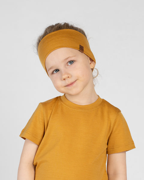 A close-up studio portrait of a young child with a slight smile, tilting her head and looking directly at the camera against a plain light grey background. She is wearing a mustard yellow short-sleeved t-shirt and a matching wide mustard yellow headband with a small brown logo tag on the side.
